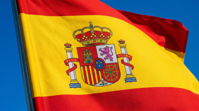 Primer plano del escudo de la bandera de España ondeando bajo un cielo azul intenso, destacando los detalles de las columnas de Hércules y la corona real