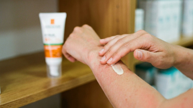 Primer plano de una mano aplicando una crema solar de textura fluida sobre un brazo con ligeras rojeces, con un envase de farmacia al fondo