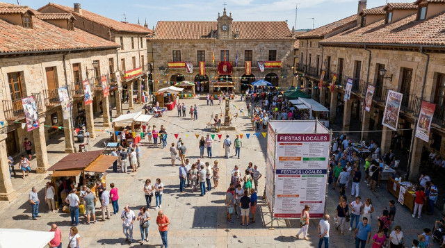 vista aérea de la plaza mayor de un pueblo español con carteles anunciando fiestas locales y gente paseando, texto 'festivos locales' visible en un panel informativo