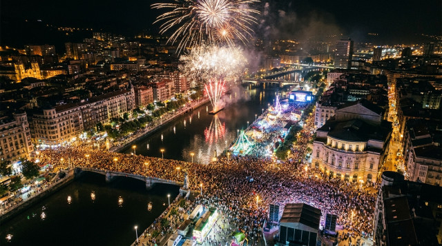 vista aérea nocturna de la zona del Arenal en Bilbao durante la Aste Nagusia con fuegos artificiales y muchedumbre en calles iluminadas