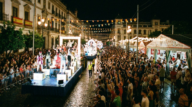 desfile nocturno por la calle principal de una ciudad española con carrozas iluminadas, público a ambos lados, banderines y una caseta de feria al fondo