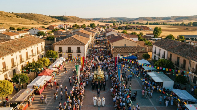imagen aérea de una romería de agosto en un pueblo español con procesión, gente en la plaza y banderolas veraniegas, luz cálida típica de tarde de verano
