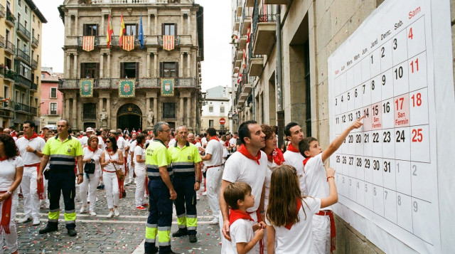 Fachada del Ayuntamiento de Pamplona con banderas, ambiente festivo urbano y gente consultando un calendario impreso que destaca 'San Fermín' como festivo local