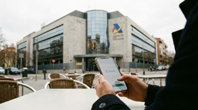 Persona utilizando un teléfono móvil para realizar una transferencia de dinero mediante Bizum con la sede de la Agencia Tributaria de fondo