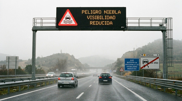 Un panel luminoso en una autovía española mostrando una de las nuevas señales de la DGT de advertencia de peligro por niebla o pérdida de visibilidad, con coches circulando de form