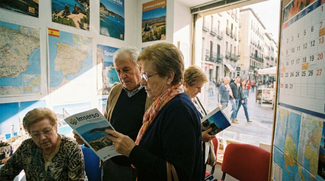 Personas mayores consultando un folleto turístico y el calendario de viajes del Imserso en una agencia de viajes